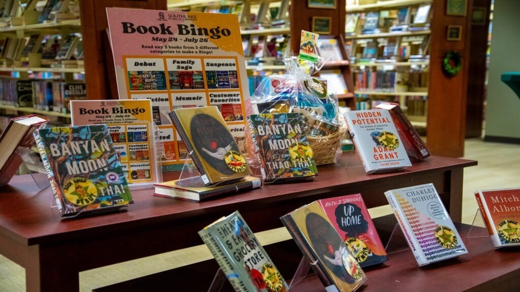 A library display of various books on wooden tables. The display includes a promotional sign for "Book Bingo" with categories like "Debut" and "Family Saga." Several book covers and a gift basket are visible amidst the backdrop of bookshelves.